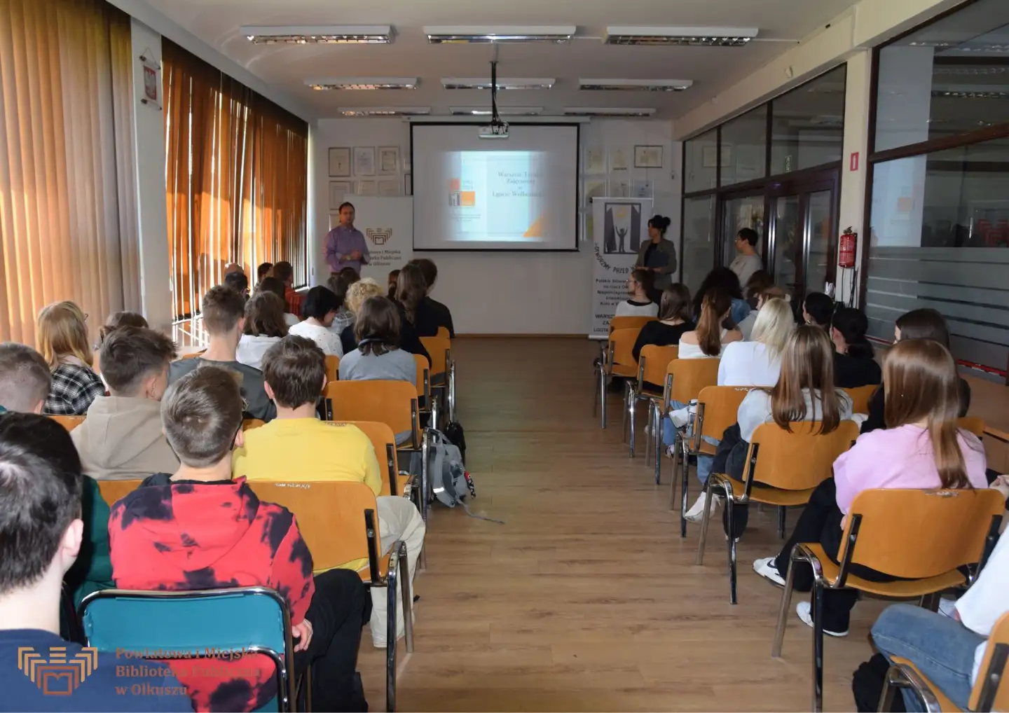 Grupa młodzieży siedzi na krzesłach tyłem do obiektywu kamery. Zgromadzeni patrzą się na dorosłego, stojącego mężczyznę obok ekranu projektora.