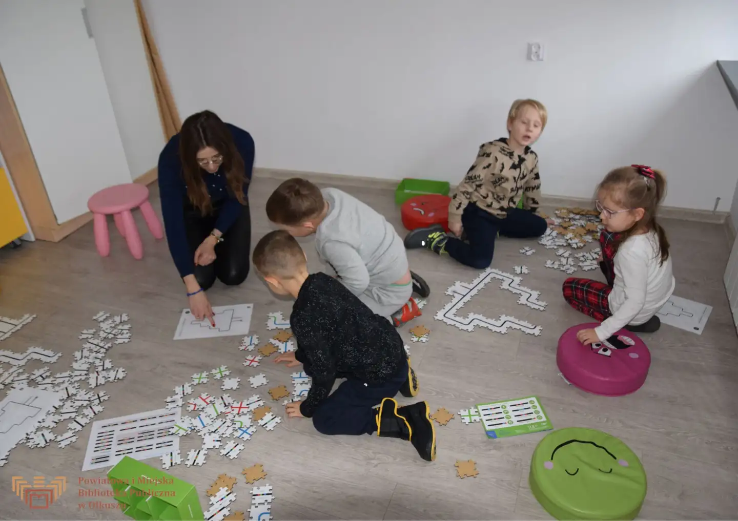 Zdjęcie zostało zrobione w sali edukacyjnej olkuskiej Biblioteki. Przedstawia kilkoro dzieci: chłopców i dziewczynki, którzy na podłodze układają puzzle - trasę dla ozobotów - w kształcie świątecznej choinki.