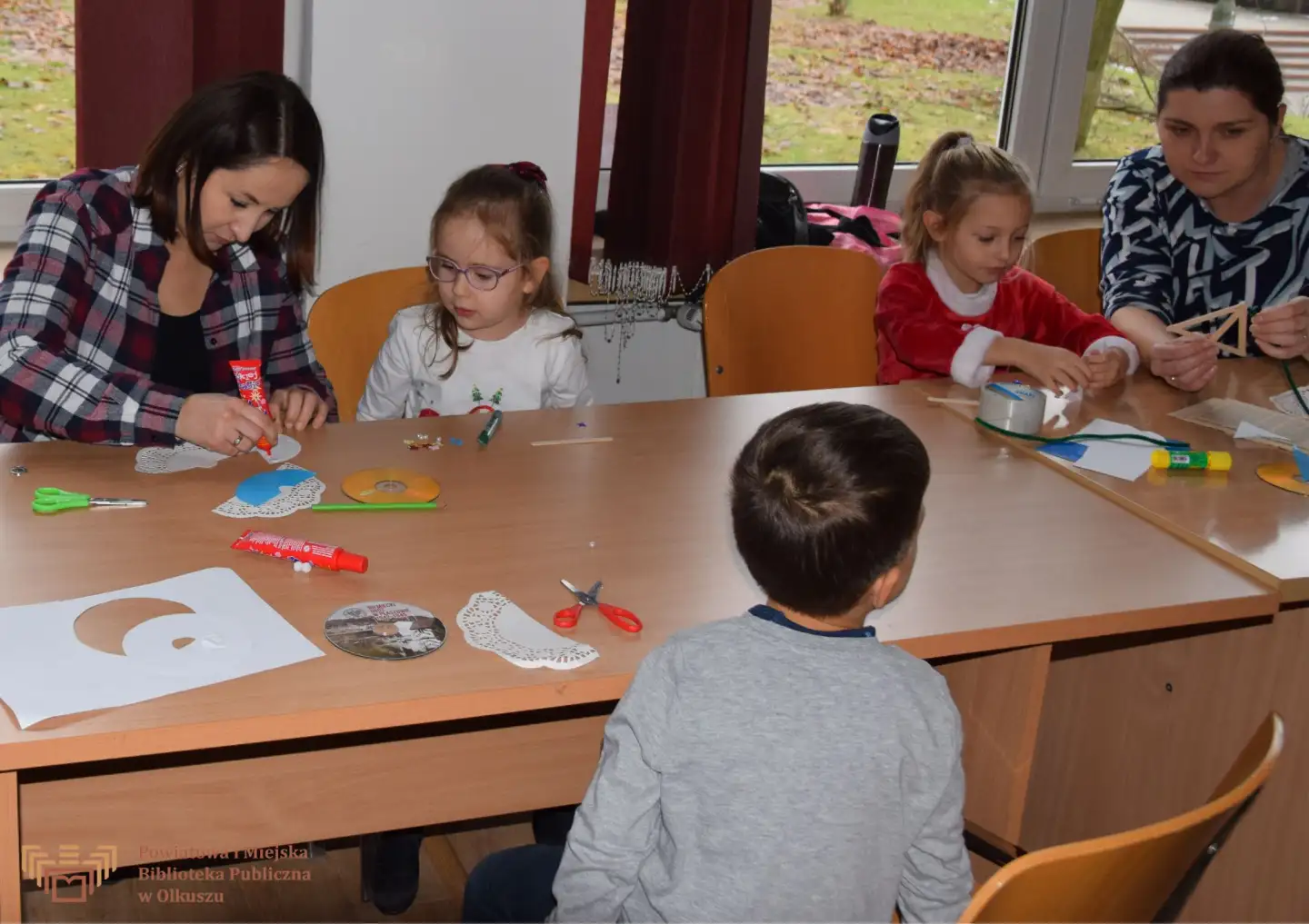 Zdjęcie zostało zrobione w Czytelni olkuskiej Biblioteki. Przedstawia grupę ludzi: dzieci i ich opiekunów, którzy siedzą przy stolikach i wykonują ozdoby świąteczne.