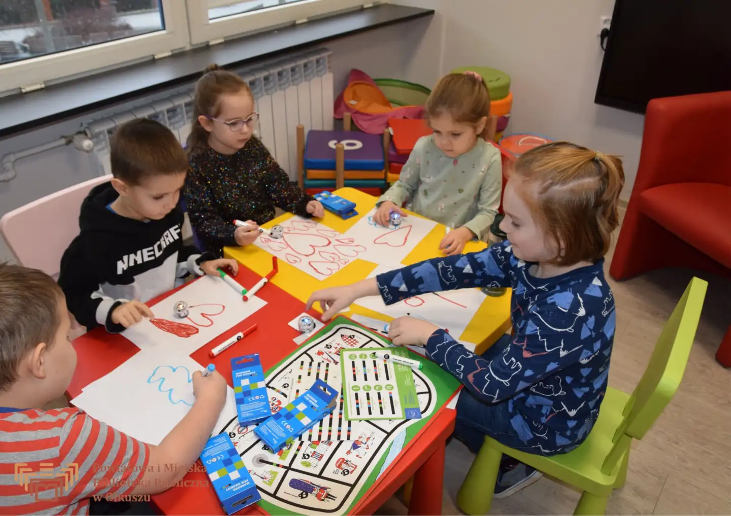 Zdjęcie przedstawia grupe dzieci siedzących przy kolorowych stolikach. Wykonują grubymi pisakami rysunki, będące trasą jazdy ozobotów.