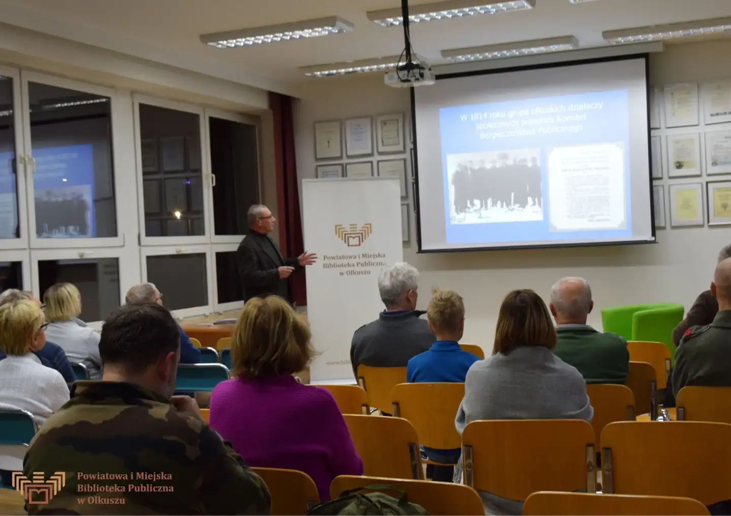 Zdjęcie przedstawia grupę osób siedzących na krzesłach w Czytelni olkuskiej Biblioteki. Są zwróceni w stronę mężczyzny prowadzącego spotkanie. Za nim na ekranie wyświetlany jest slajd prezentacji.