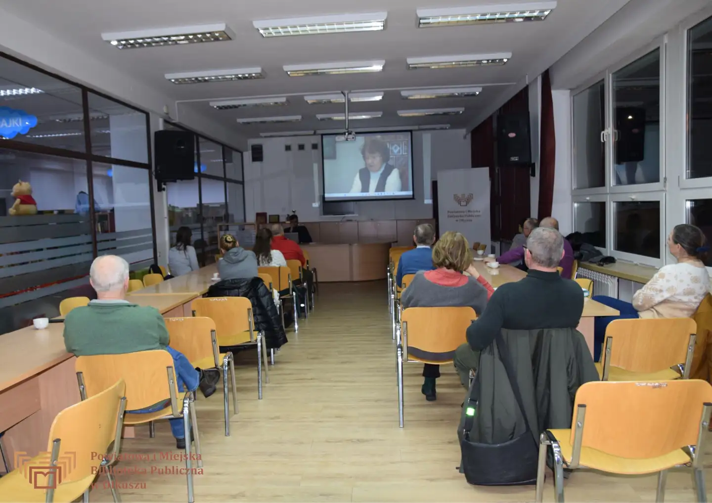 Zdjęcie zostało zrobione w Czytelni olkuskiej Biblioteki. Przedstawia grupę dorosłych ludzi siedzących przy stolikach. Są zwróceni w stronę mężczyzny prowadzącego wykład. Nad nim, na dużym ekranie, wyświetlany jest film.