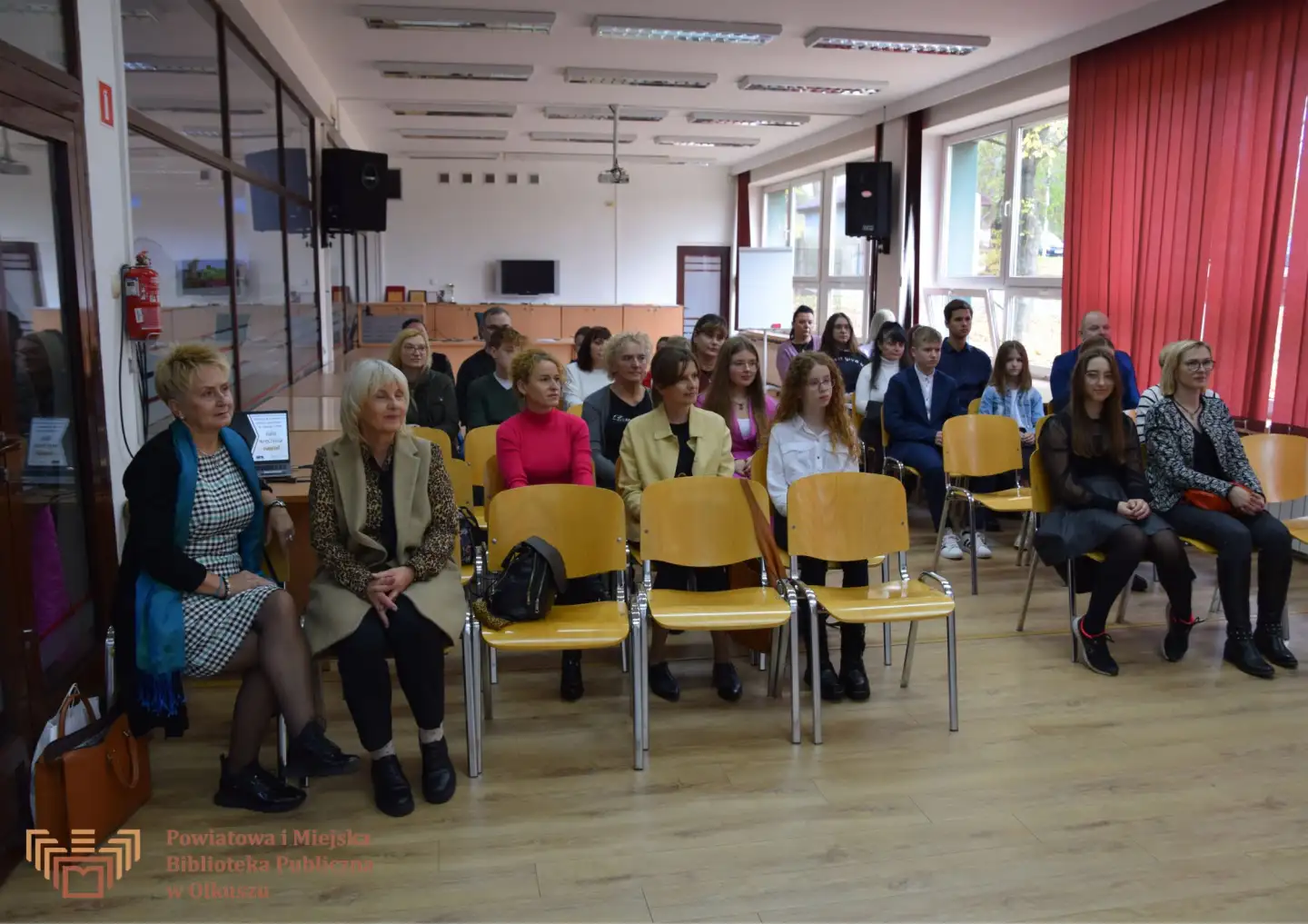 Zdjęcie zostało zrobione w Czytelni olkuskiej Biblioteki. Przedstawia grupę dzieci, młodzieży oraz ich opiekunów siedzących na krzesłach.