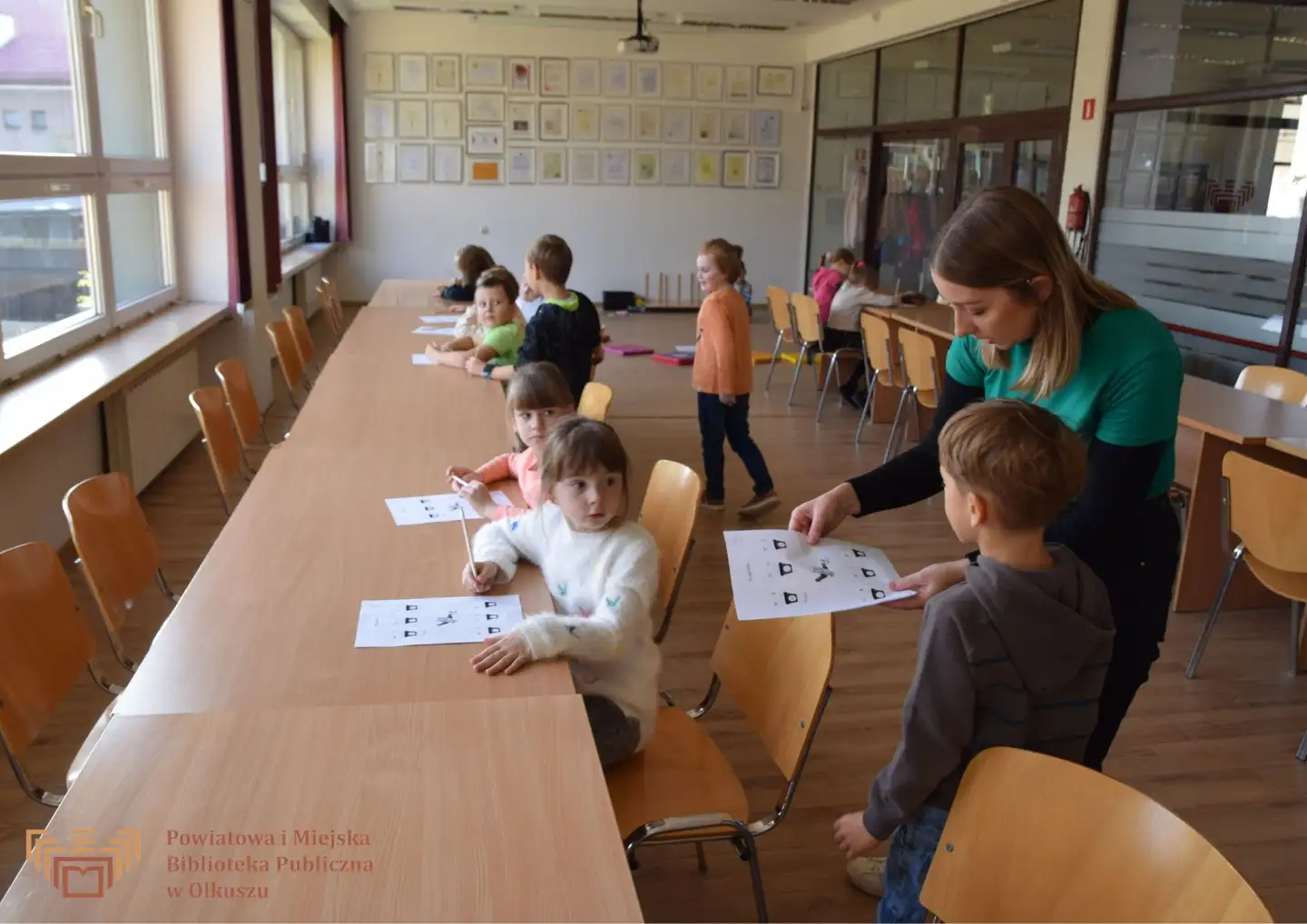 Zdjęcie zostało zrobione w Czytelni olkuskiej Biblioteki. Przedstawia dzieci siedzące przy stolikach. Rozwiązują zadania, które daje im Pani w zielonym podkoszulku.