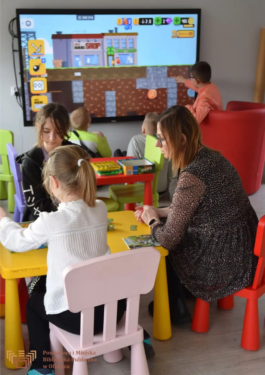 Zdjęcie zrobione w trakcie zajęć GraiBoty - gry planszowe i roboty. Dorosła kobieta i dwójka dziewczyn siedzi przy stoliczku. W tle widać chłopców grających na tablicy interaktywnej.