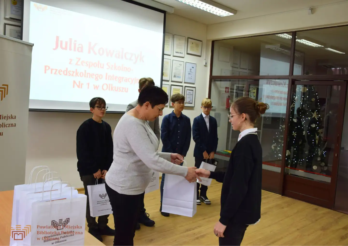 Zdjęcie zostało zrobione w Czytelni Biblioteki. Przedstawia Panią Dyrektor wręczającą nagrodę jednej z laureatek. W tle widać grupę pozostałych laureatów.