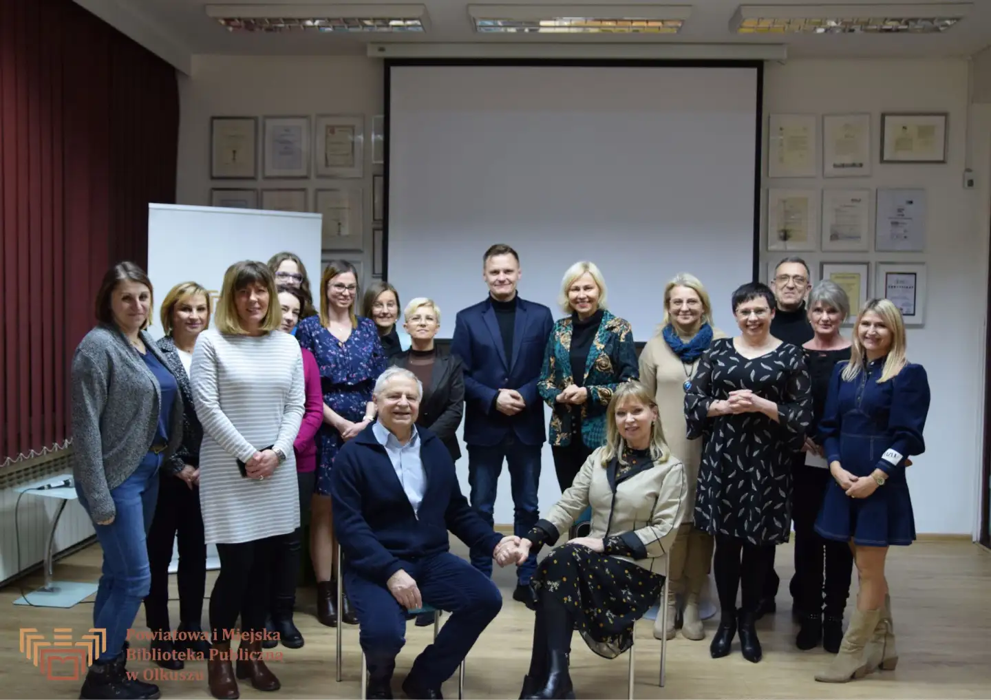 Zdjęcie zostało zrobione w Czytelni olkuskiej Biblioteki. Przedstawia grupę uczestników spotkania z aktorami. Pośrodku na krzesłach siedzą występujący aktorzy: Dorota Segda i Jacek Romanowski. Trzymają się za ręce.