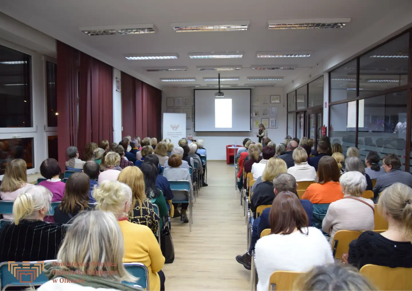 Zdjęcie zostało zrobione w Czytelni olkuskiej Biblioteki. Przedstawia siedzących na krzesłach uczestników spotkania z aktorami: Dorotą Segdą i Jackiem Romanowskim.