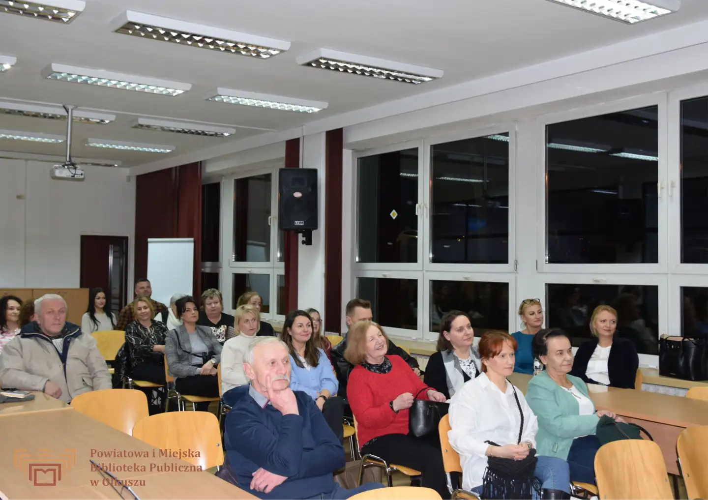 Zdjęcie przedstawia grupę ludzi siedzących w dużej, bibliotecznej sali.