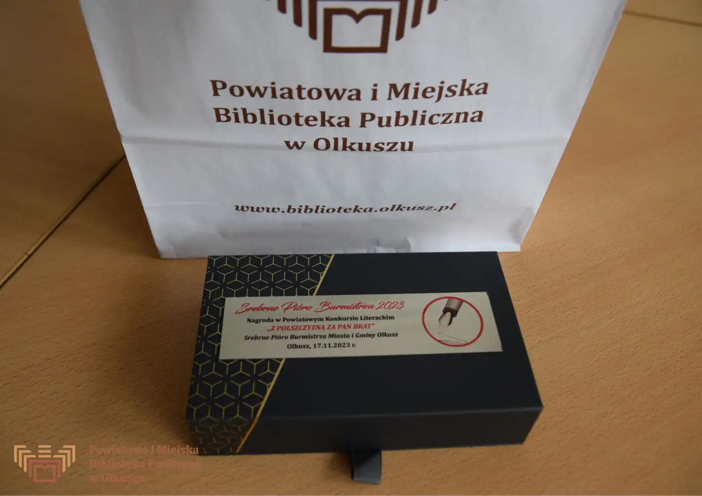 Zdjęcie przedstawia leżący na stoliku czarno-złoty futerał z naklejoną plakietką z napisem Srebrne Pióro Burmistrza 2023. Nagroda w Powiatowym Konkursie Literackim "Z polszczyzną za Pan brat" o Srebrne Pióro Burmistrza Miasta i Gminy Olkusz. Obok stoi biała torba z logo i napisem: Powiatowa i Miejska Biblioteka Publiczna w Olkuszu.
