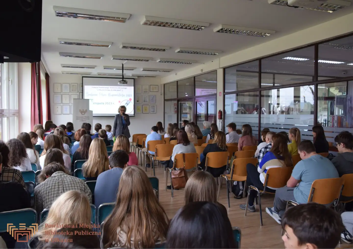 Zdjęcie przedstawia grupę ludzi siedzących na krzesłach w Czytelni olkuskiej Biblioteki. Są oni zwróceni w stronę kobiety stojącej na środku sali i mówiącej do mikrofonu. Za nią stoi biały baner z logo i napisem Powiatowa i Miejska Biblioteka Publiczna w Olkuszu.