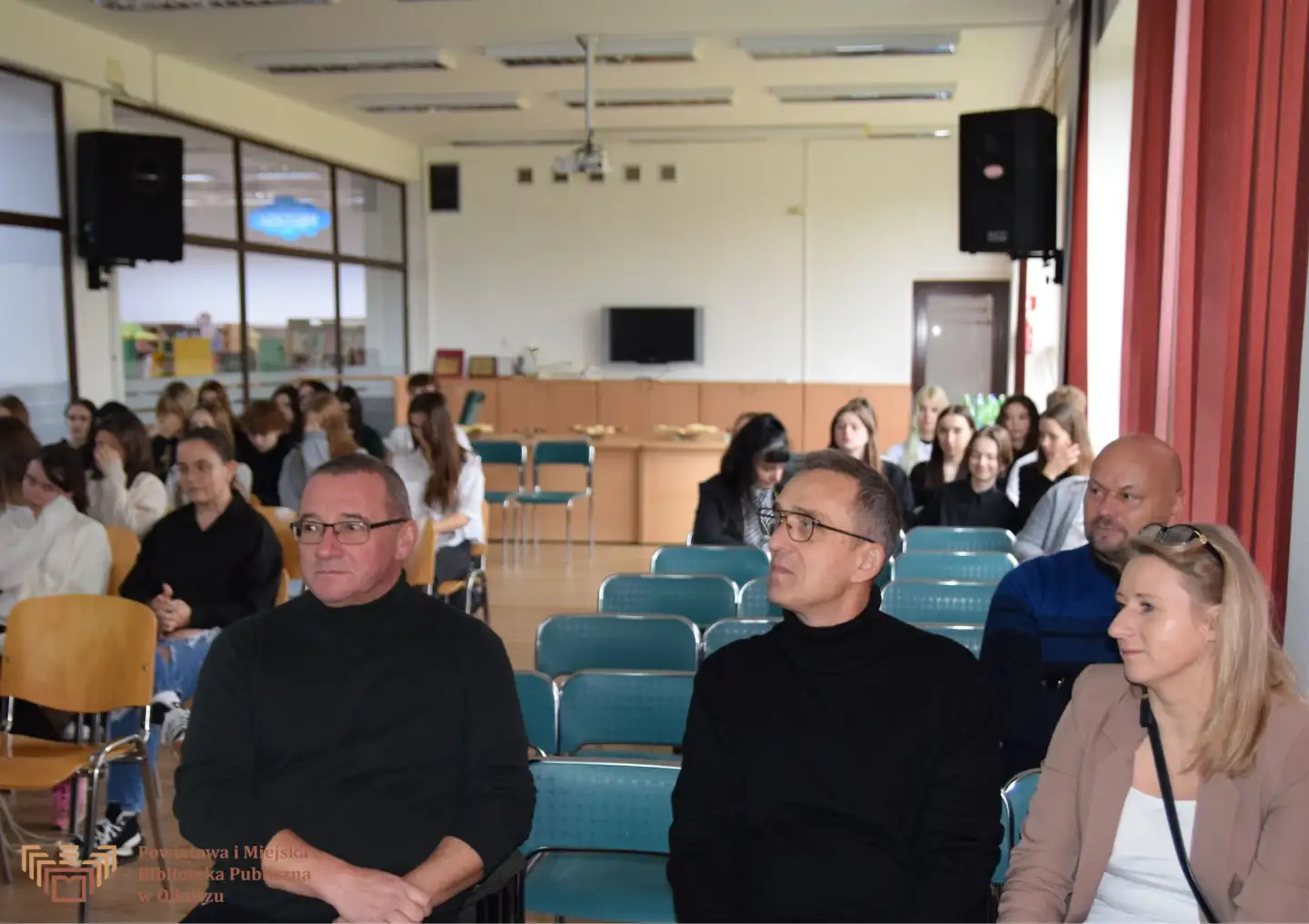 Zdjęcie przedstawia grupę ludzi siedzących na krzesłach w Czytelni olkuskiej Biblioteki. Na pierwszym planie widzimy dwóch mężczyzn w okularach i czarnych golfach oraz kobietę w beżowej marynarce.