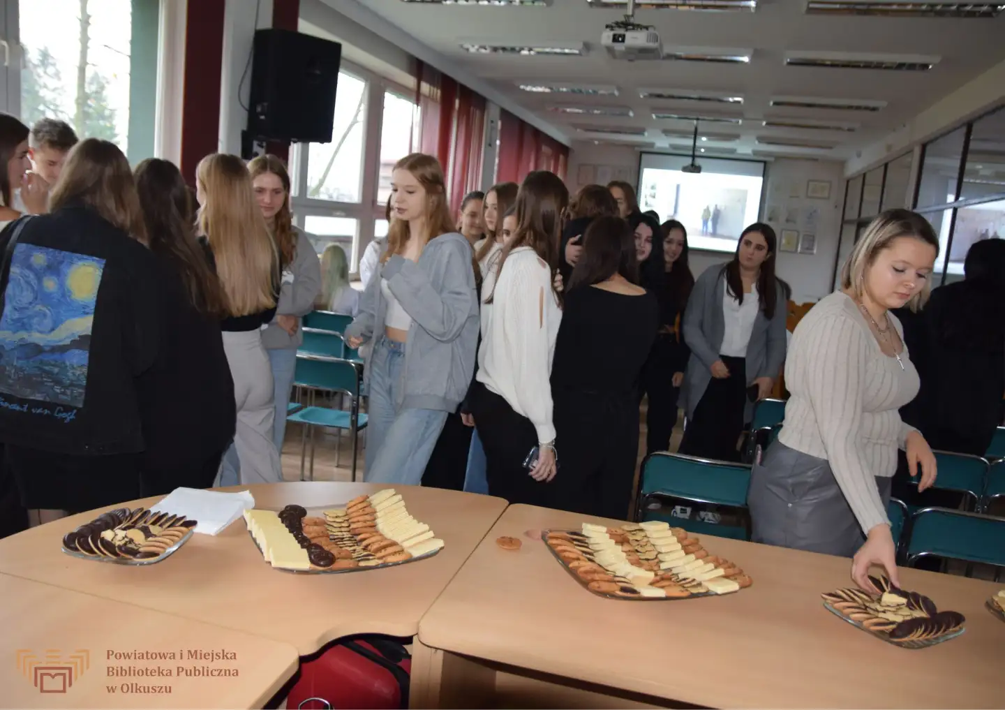 Zdjęcie przedstawia grupę ludzi stojących obok stolików w Czytelni olkuskiej Biblioteki. Na stolikach leżą patery z ciastkami.