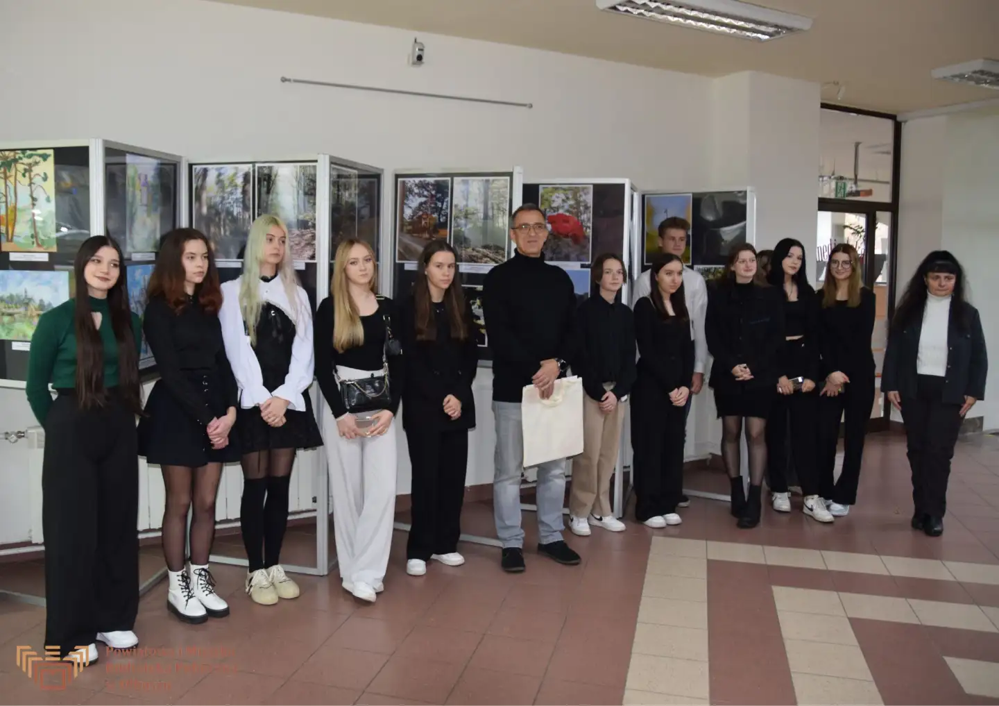 Zdjęcie przedstawia grupę młodzieży wraz z dwójką opiekunów pozujących do zdjęcia na tle wystawy z pracami uczniów.
